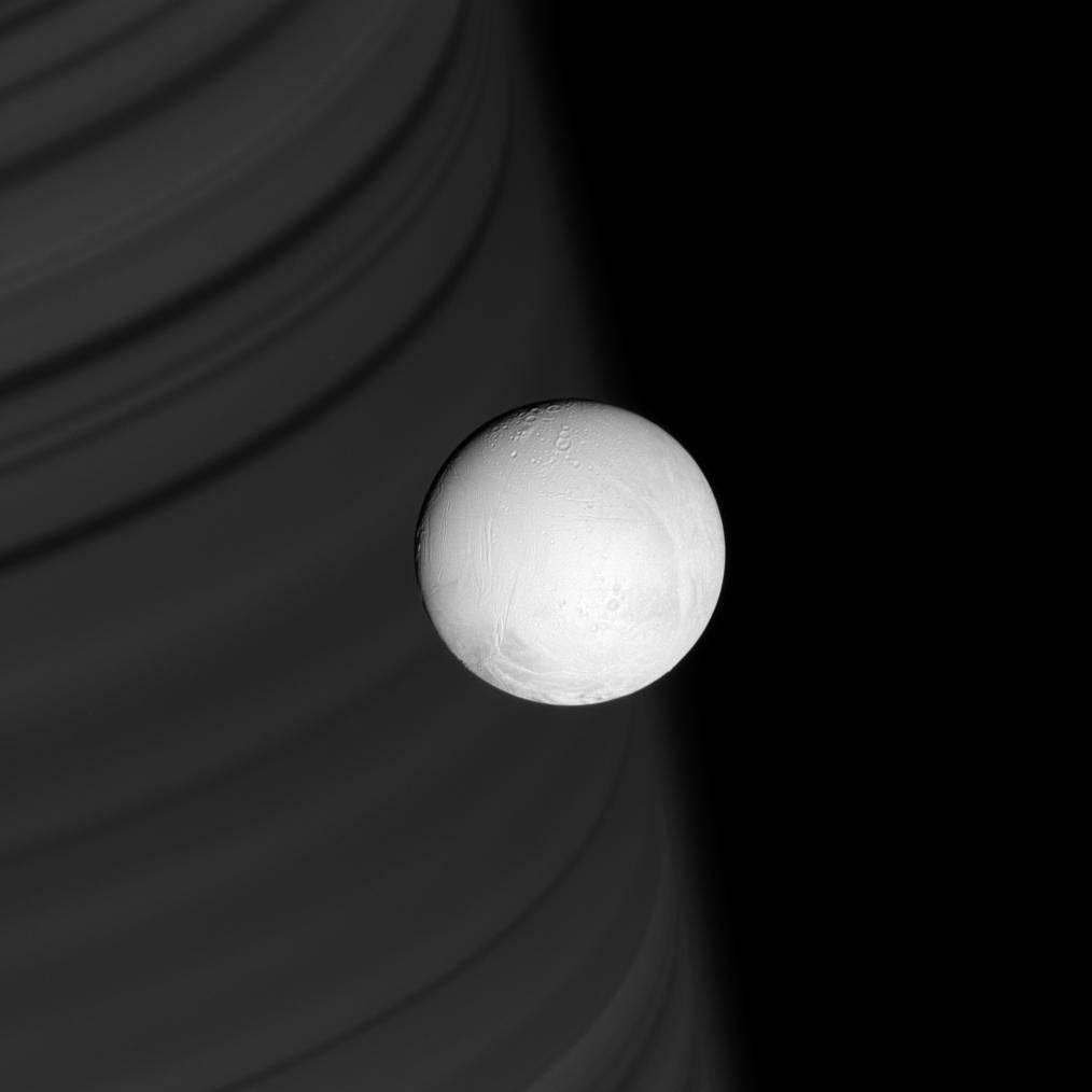 Ring shadows line the face of distant Saturn, providing a backdrop for the brilliant, white sphere of Enceladus.