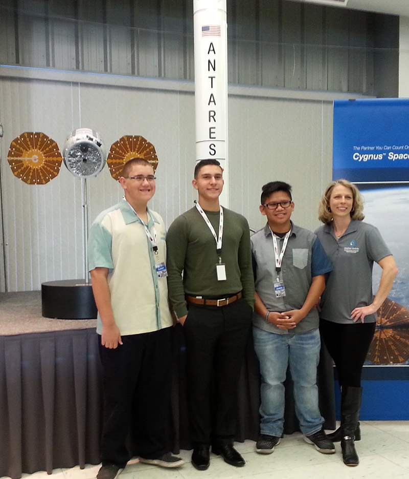 Devin (second from left) poses with collaborators in front of models of the Antares rocket and Cygnus capsule.