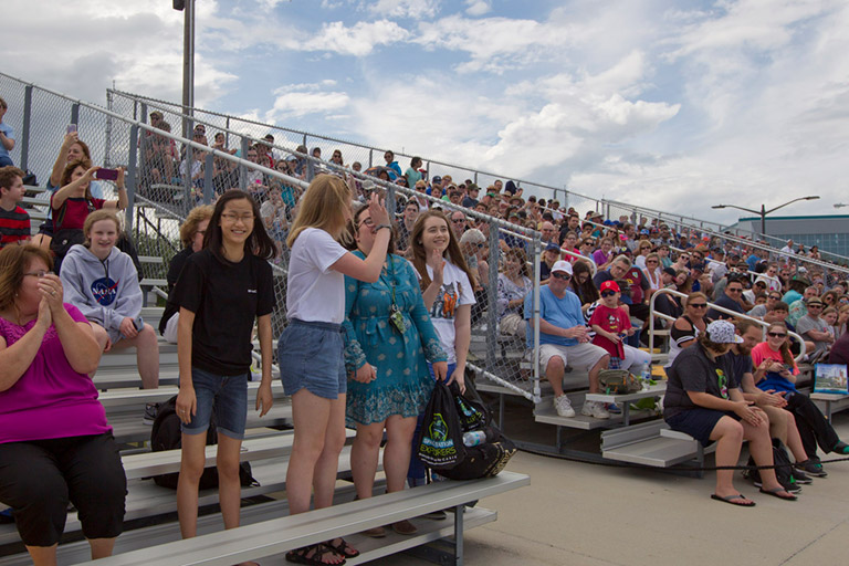 16. As the launch window neared, students and their mentors, families, and friends gathered on the bleachers at the Banana Creek viewing area next to the Apollo Saturn V building. An announcer invited each team to stand for applause.