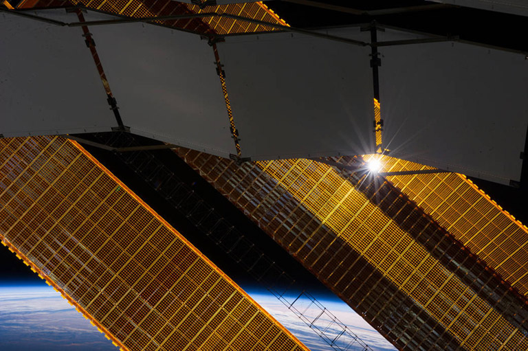 ISS038 E 024901 (2 Jan. 2014) A tiny representation of the sun sneaks through between a truss based radiator panel and a primary solar array panel on the Earth orbiting International Space Station in this photograph taken by one of the Expedition 38 crew members on Jan. 2, 2014. Clouds over Earth and the blackness of space share the background scene.