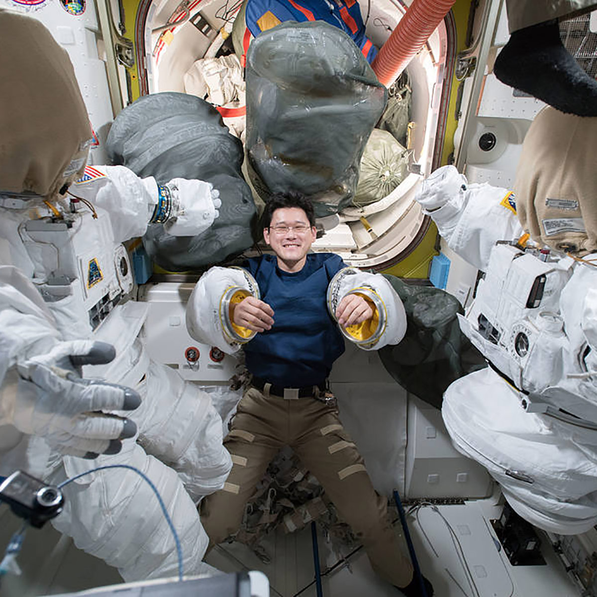 iss054e014016 (Jan. 2, 2018) Astronaut Norishige Kanai of the Japan Aerospace Exploration Agency tries on a pair of spacesuit sleeves inside the Quest airlock. To either side of Kanai are the U.S. spacesuits that were being readied for a pair of robotics maintenance spacewalks.