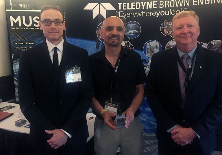 Paul Galloway from Teledyne Brown Engineering (right) with RPI researchers Joe Adam (left) and Amir Hirsa (middle), who is holding a Ring Sheared Drop system test cell, at the 2022 ISS Research and Development Conference.