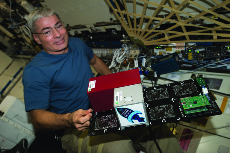 This photo from December 2017 shows Mark Vande Hei attaching modular CubeLabs (boxes) onto a Tangolab card (large panel) onboard the ISS. The housing for Team Orions experiment looks just like the silver CubeLab decorated with the rocket shaped <em>Go For Launch!< em> sticker.” width=”768″ height=”511″></div><figcaption class=