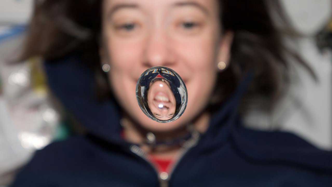 megan-mcarthur-water-bubble STS-125 Mission Specialist Megan McArthur posing for a photo on the middeck. A water bubble, showing a reflection of McArthur, floats in front of her.